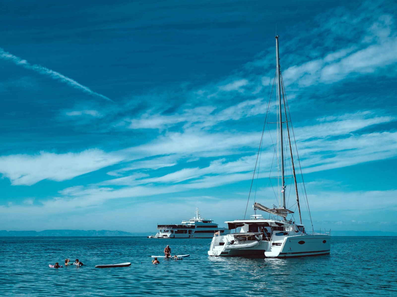 A catamaran at anchor on calm Mediterranean water, with guests swimming and paddleboarding beside it